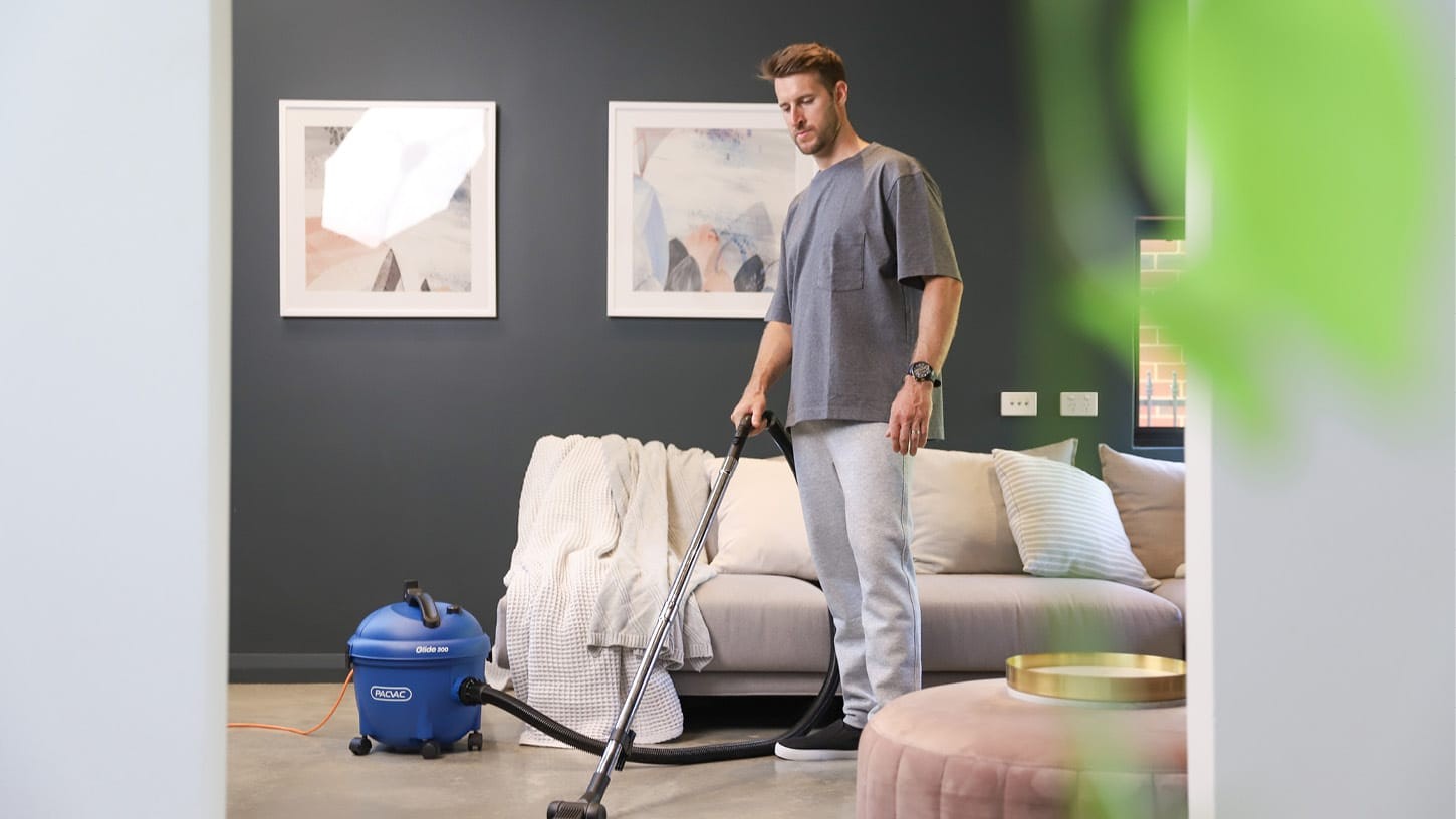 Male in comfortable clothing using a canister vacuum cleaner to vacuum the shiny floor of a living room, among modern looking furniture.