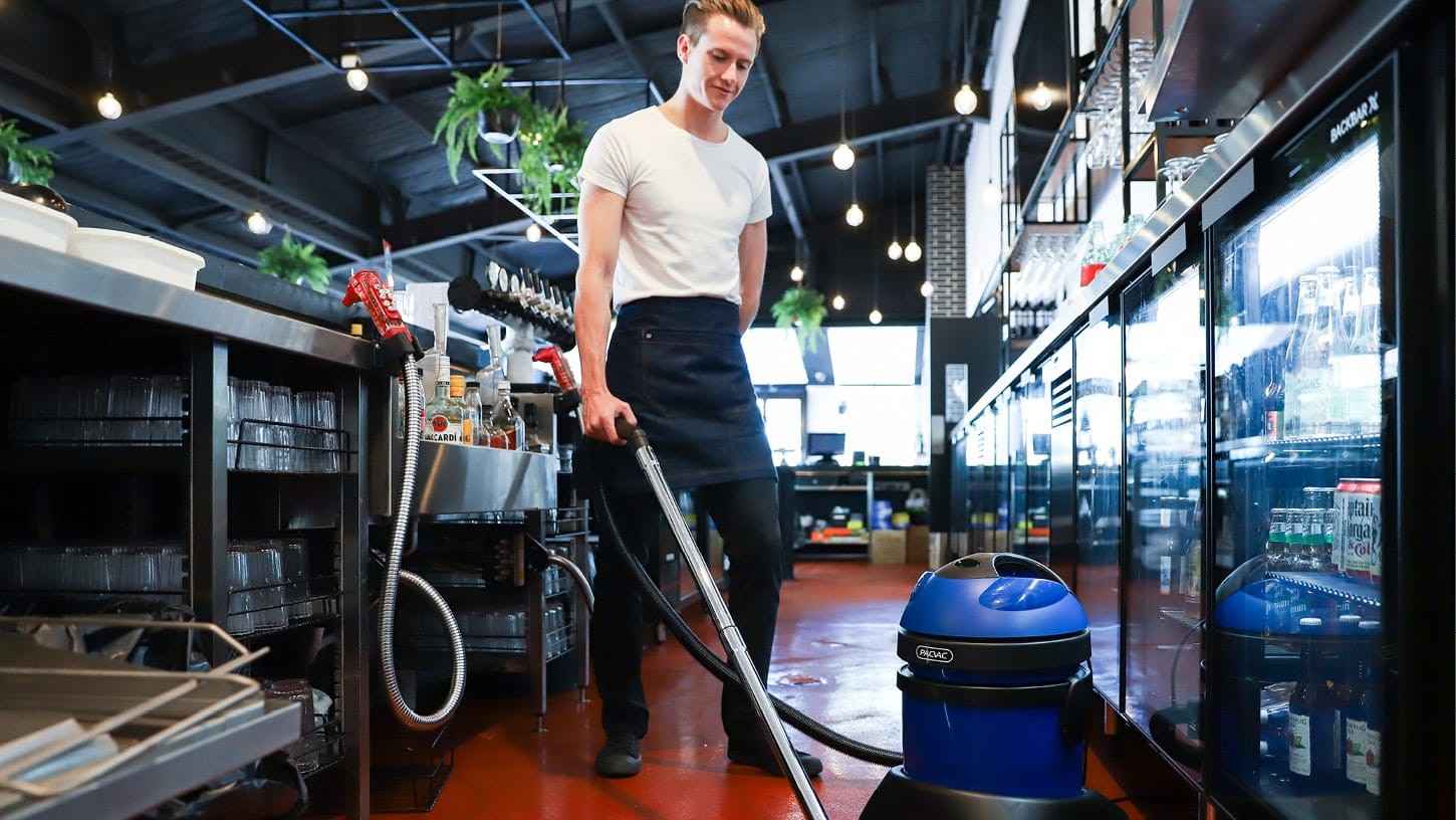 A professional wet and dry vacuum is being used by a hospitality worker to vacuum the shiny red floor of a bar, with shelves containing glassware and alcohol.