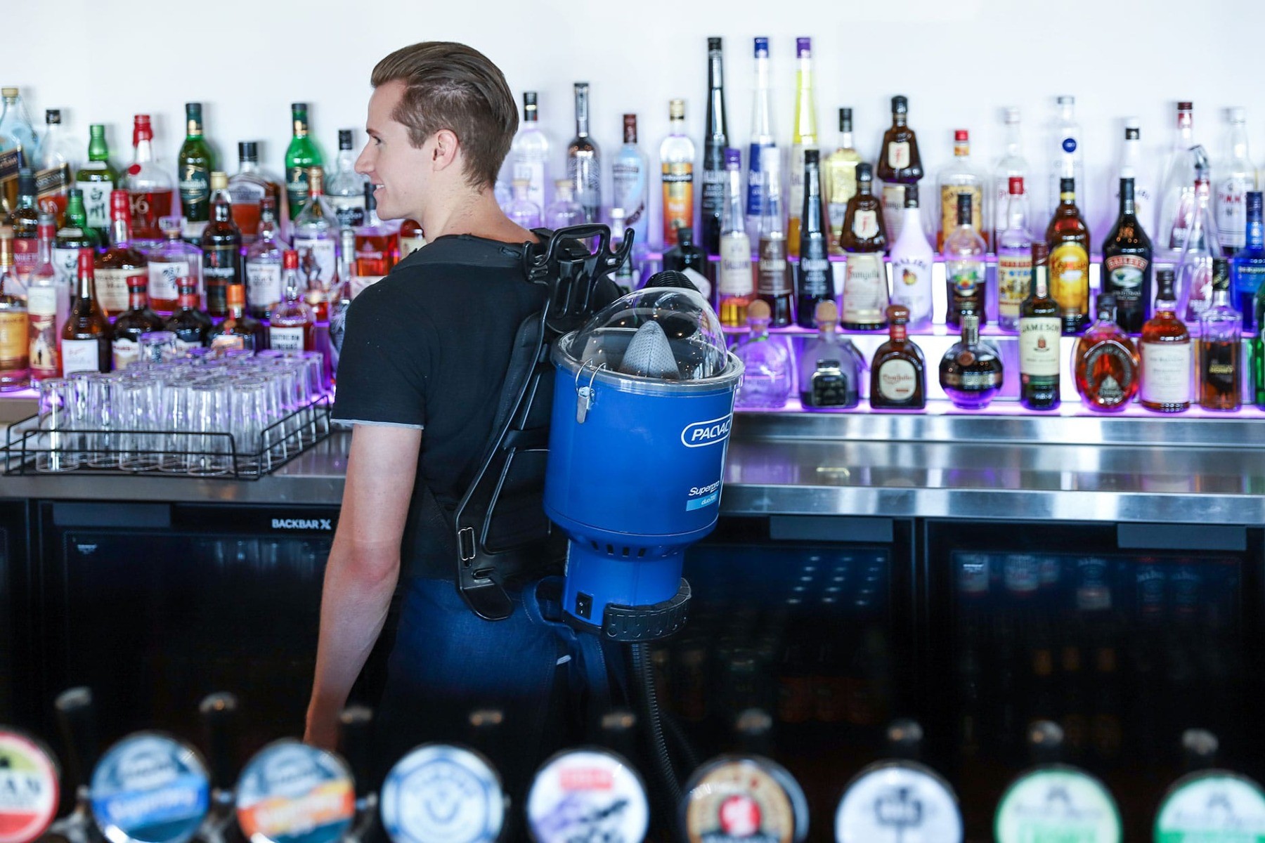 Male in a bar with lots of liquor and glassware using a water-proof backpack vacuum.