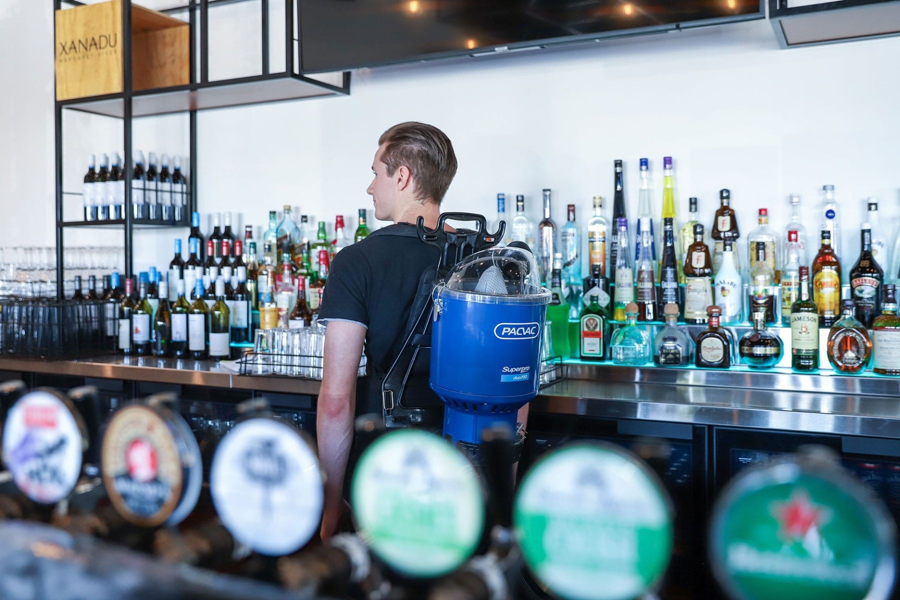 Young male behind a well stocked bar with lots of liquor wearing a Superpro duo 700 backpack vacuum.