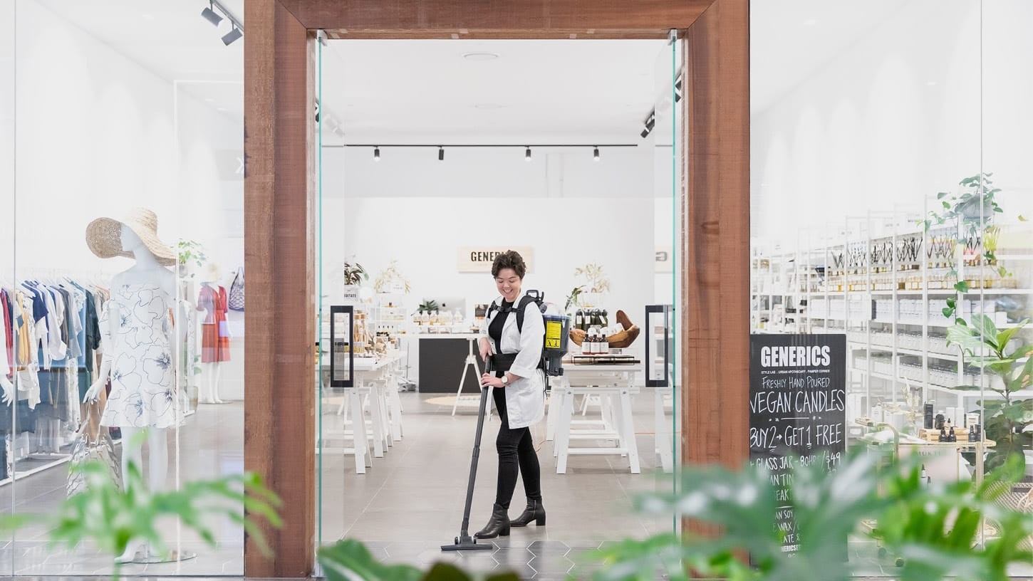 Photo of the storefront of a boutique retail shop called Generics, with large glass windows and shop assistant smiling as she's vacuuming the floor with a backpack vacuum.